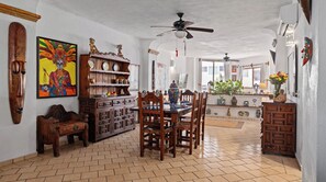 Un coin repas avec une table en bois, des chaises et un ventilateur de plafond. On y trouve des objets décoratifs sur des étagères et un tableau au mur.