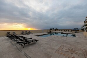A rooftop pool area with sun loungers and a swimming pool, surrounded by a low wall and palm trees.