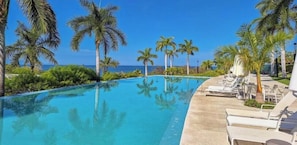 A clear blue pool with lounge chairs and palm trees, overlooking the ocean.