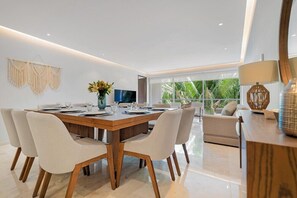 A modern dining area with a wooden table, white upholstered chairs, a vase with flowers, and a television. - Punta de Mita 4BR/4BA Condo w Pool, Beach & Gym (Corral del Risco)
