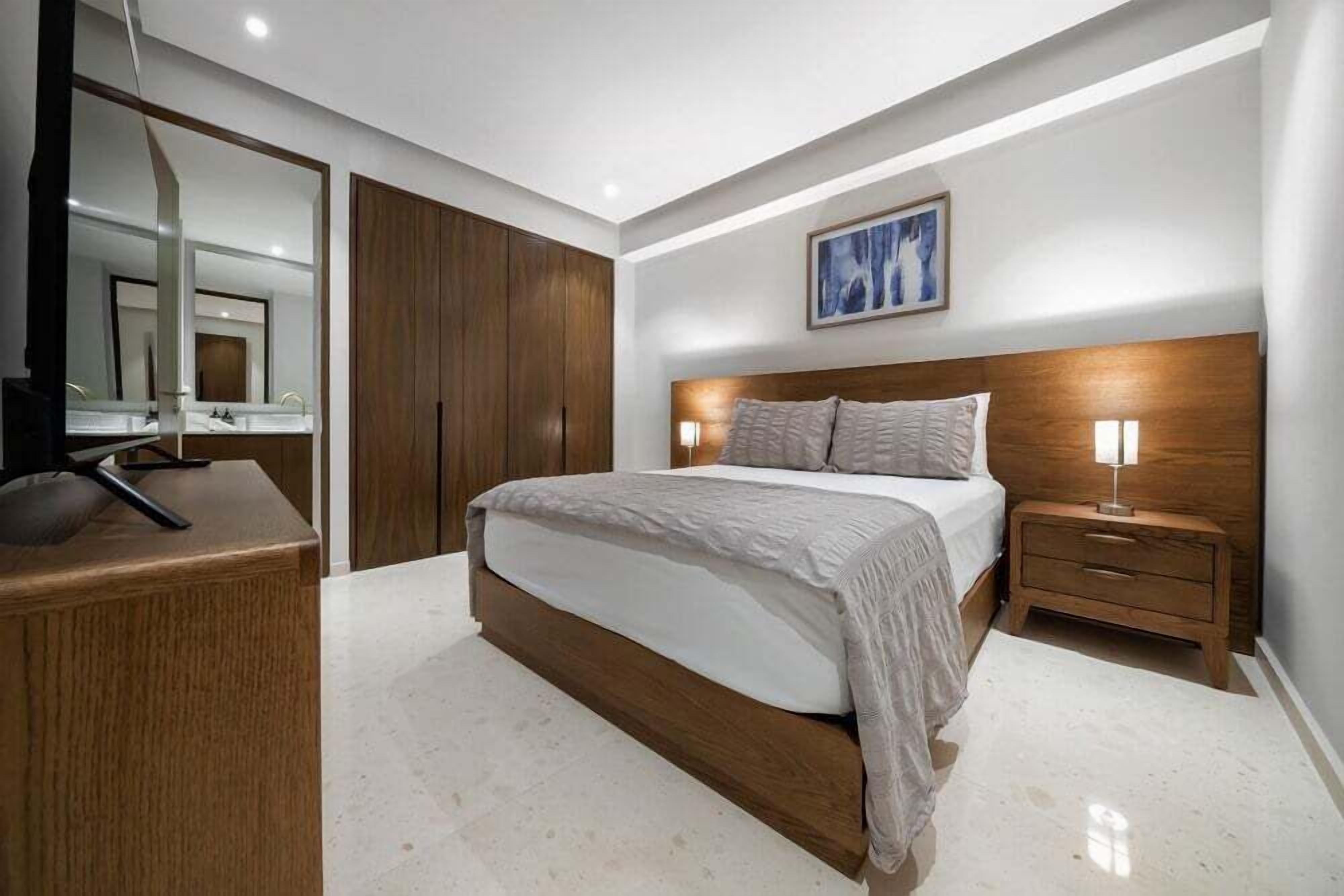 Ein modernes Schlafzimmer mit einem großen Bett, einem Holzschrank, einem Nachttisch mit Lampe und einem Spiegel.