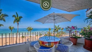 A balcony with a round table and chairs, overlooking a beach with palm trees and the ocean.