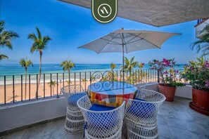 Un balcón con mesa y sillas, con vistas a una playa con palmeras y el océano.