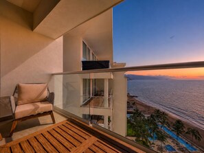 Een balkon met uitzicht op zee bij zonsondergang, een houten terras en een zithoek met een stoel en een tafel.