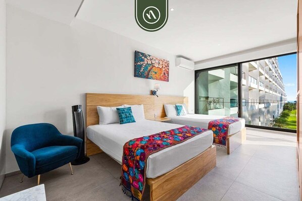 Quarto de hotel moderno com uma cama grande, uma poltrona azul e vista para um edifício através de grandes janelas.