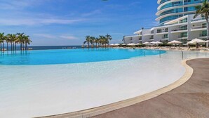 Una piscina grande y despejada con borde curvo, rodeada de palmeras y un edificio moderno de hotel con varios balcones.