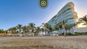 Un hotel frente a la playa con varios balcones, palmeras y sombrillas.