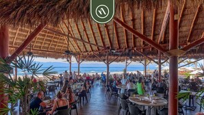 Un área de comedor al aire libre con techos de paja y piso de madera, con vista a la playa.