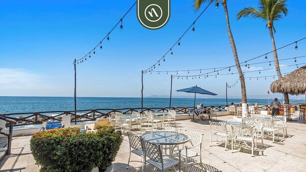 An outdoor seating area with tables and chairs, a thatched-roof structure, and a view of the sea.