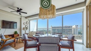 A spacious living room with a round dining table, multiple chairs, a ceiling fan, and large windows offering a city view.