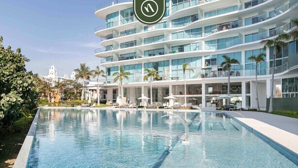 A modern hotel with a large swimming pool, multiple balconies, and palm trees.