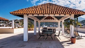 A covered outdoor seating area with a tiled roof, surrounded by potted plants, overlooking a residential area. - 206-VistadelSol | 2BR/2BA Ocean View, Beachfront (Puerto Vallarta)