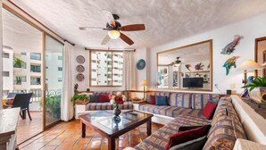 Ein geräumiges Wohnzimmer mit einer großen Couch, einem Couchtisch und einem Deckenventilator. Es gibt einen Balkon mit Tisch und Stühlen.