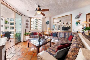 A spacious living room with a large sofa, a coffee table, and a ceiling fan. There is a balcony with a table and chairs outside.