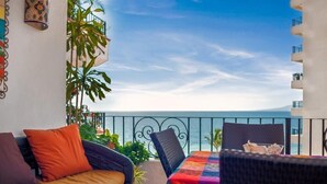 Ein Balkon mit einer Couch und Stühlen, einem Tisch mit bunter Tischdecke und Blick auf das Meer.