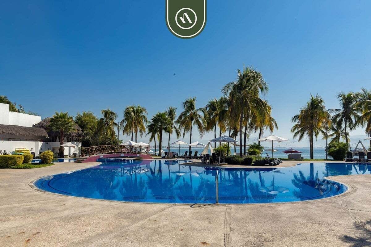 A large swimming pool surrounded by palm trees and lounge chairs, with a view of the ocean.
