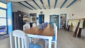 Un comedor con una mesa de madera y sillas blancas, techo azul y una cocina al fondo.