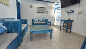 Una habitación con un sofá azul, una mesa de centro de madera, un televisor montado en la pared y un ventilador de techo.