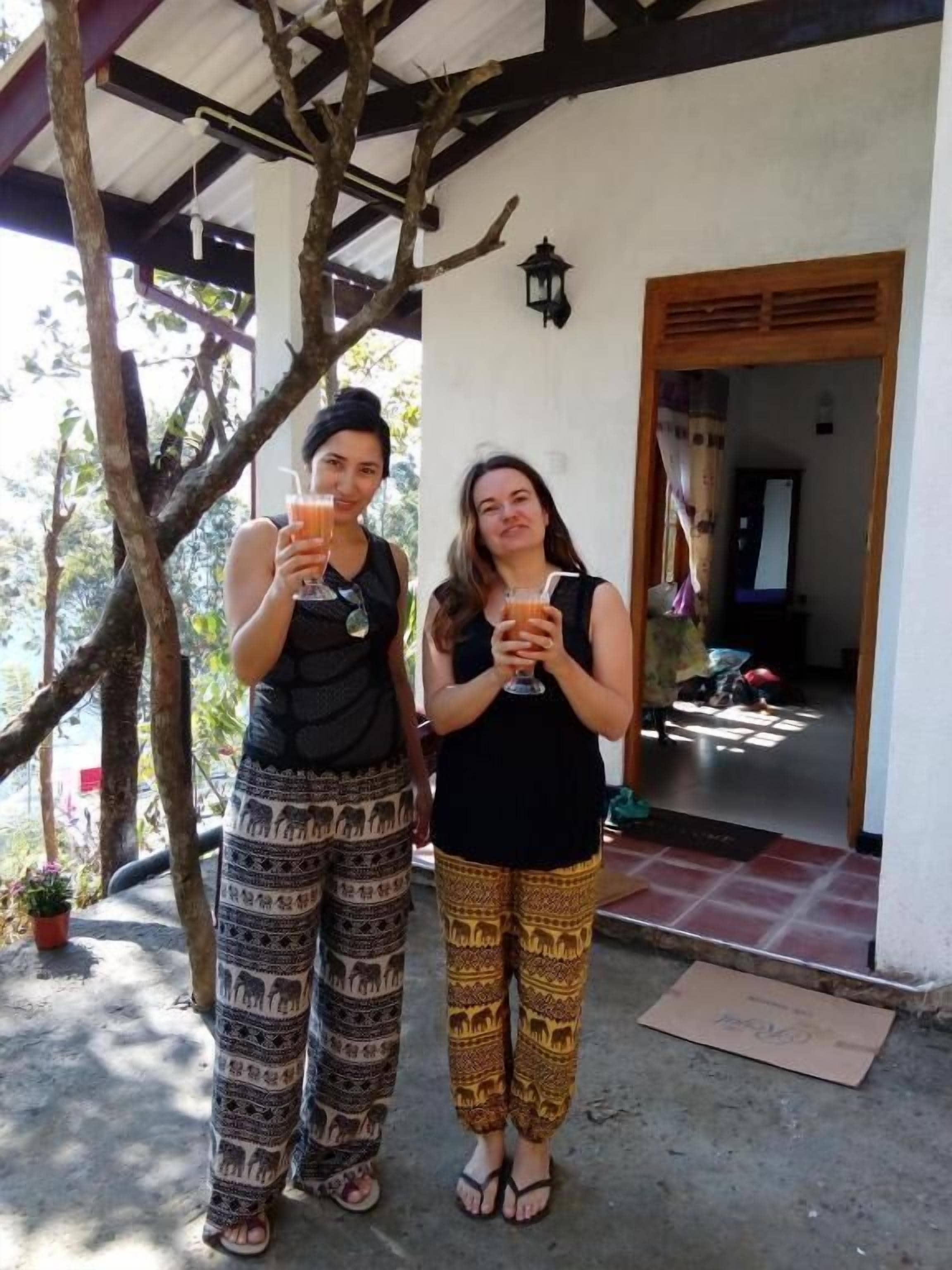 Dos personas de pie frente a un edificio, con un árbol y una puerta a la vista al fondo.