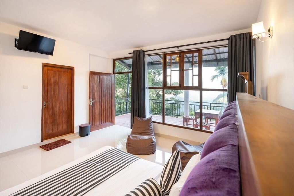 A modern hotel room with a large window, a flat-screen TV mounted on the wall, a wooden door, and a balcony with outdoor furniture.