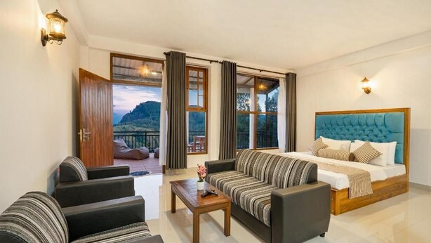 A hotel room with a bed, a sofa, a coffee table, and a view of a mountainous landscape through large windows. - Ella Ostella (Ella)