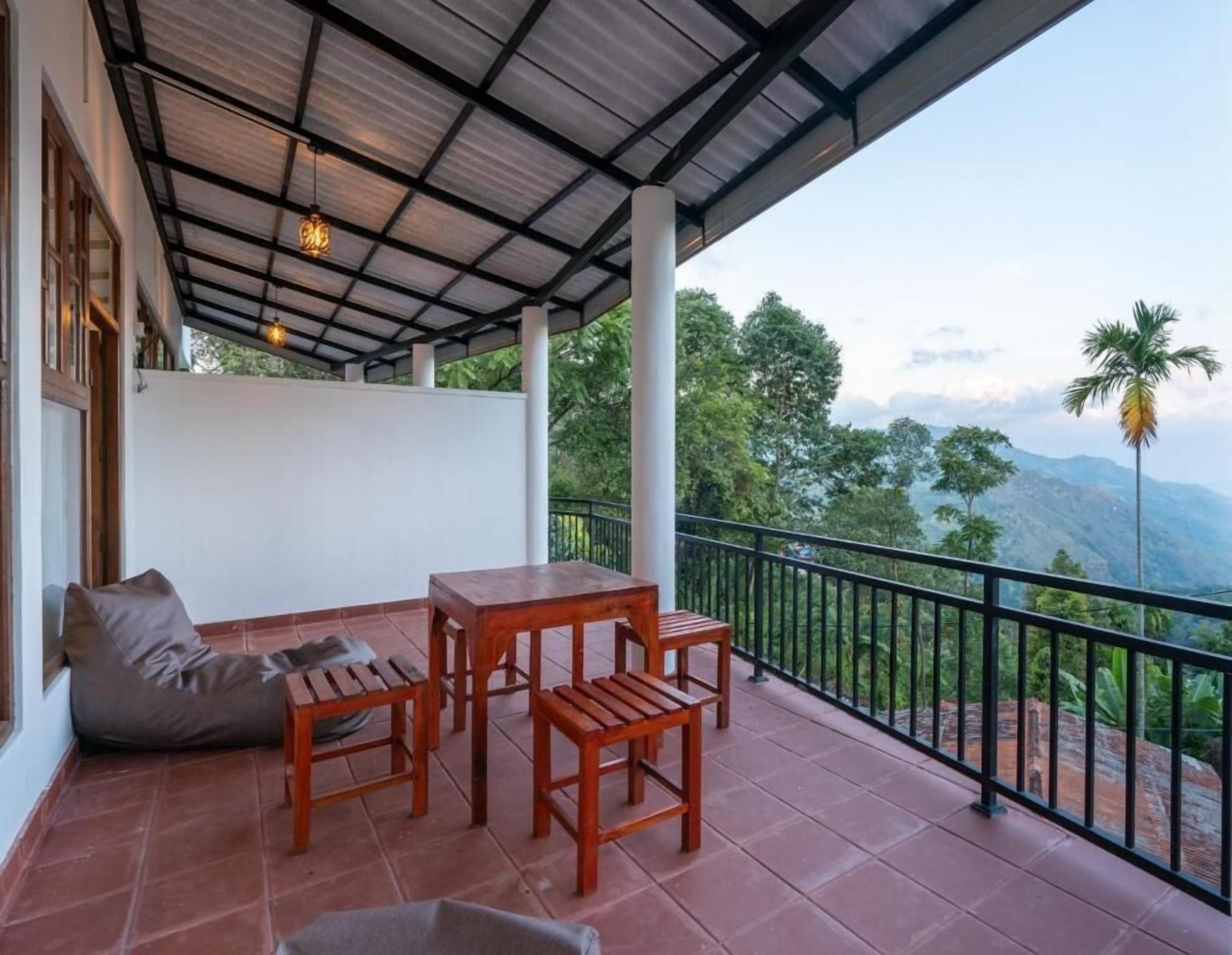 A balcony with a wooden table and chairs, a cushioned seat, and a view of the surrounding greenery and mountains.