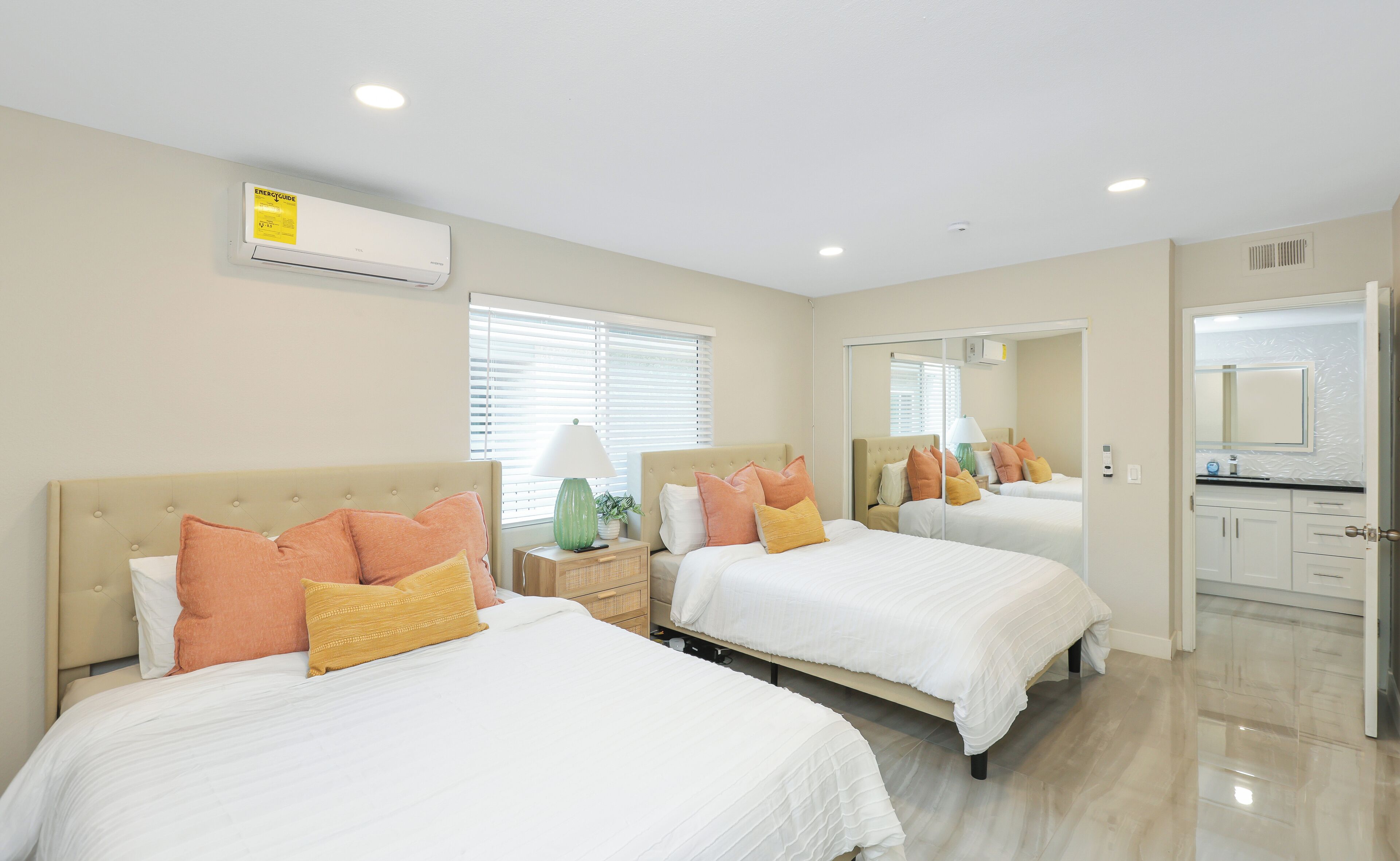 A hotel room with two beds, a mirror, a window with blinds, and a bathroom visible through an open door.