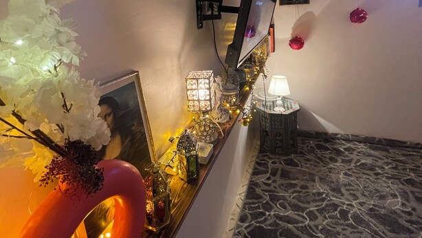 A room with a mounted TV, a bed with a patterned bedspread, a small table with decorative items, and wall-mounted picture frames.