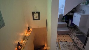 A narrow hallway with a tiled floor, a framed picture on the wall, and a staircase leading to a bathroom with a sink and shelves.