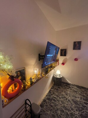 Een kamer met een wandgemonteerde televisie waarop 'HOMELAND' te zien is, een decoratieve plank met verlichting en decoraties, een kamerbreed tapijt met patroon, en een kunstwerk in een lijst aan de muur.
