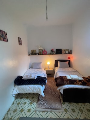 A small, clean room with two beds, a nightstand with a lamp, a shelf with various items, and a patterned floor.