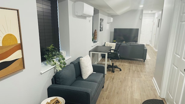A modern living room with a grey sofa, a dining table, and a TV in the background. - Exquisite private 1-bedroom apartment in delightful Newark with modern amenities (Newark)