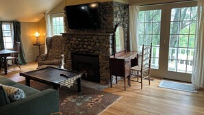 A cozy living room with a stone fireplace, a television mounted above it, and a large window with curtains. - Countryside Cottage with Sauna and Fireplace (Berlin)