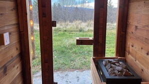 A wooden cabin with a view of a grassy field and mountains. There is a wooden bench and a fireplace with rocks. - Countryside Cottage with Sauna and Fireplace (Berlin)