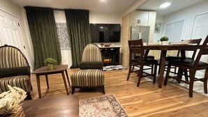 A cozy living room with striped armchairs, a wooden coffee table, and a patterned rug. A fireplace is visible in the background, and a dining area with a wooden table and chairs is adjacent to the living space.