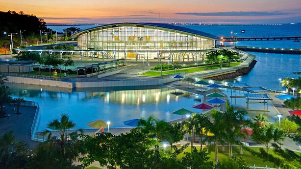A modern building with a glass facade, surrounded by a water body and a park area with umbrellas.