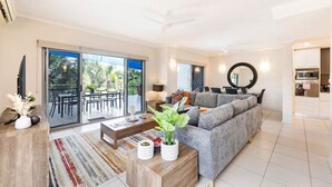 A modern living room with a grey sofa, a coffee table, and a TV. There's a balcony with outdoor furniture and a view of greenery.
