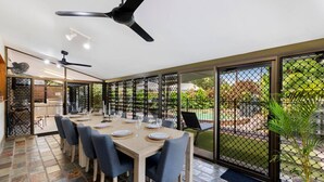 Un coin repas avec une table dressée pour six personnes, un ventilateur de plafond et une vue sur un jardin, visible à travers une porte vitrée.