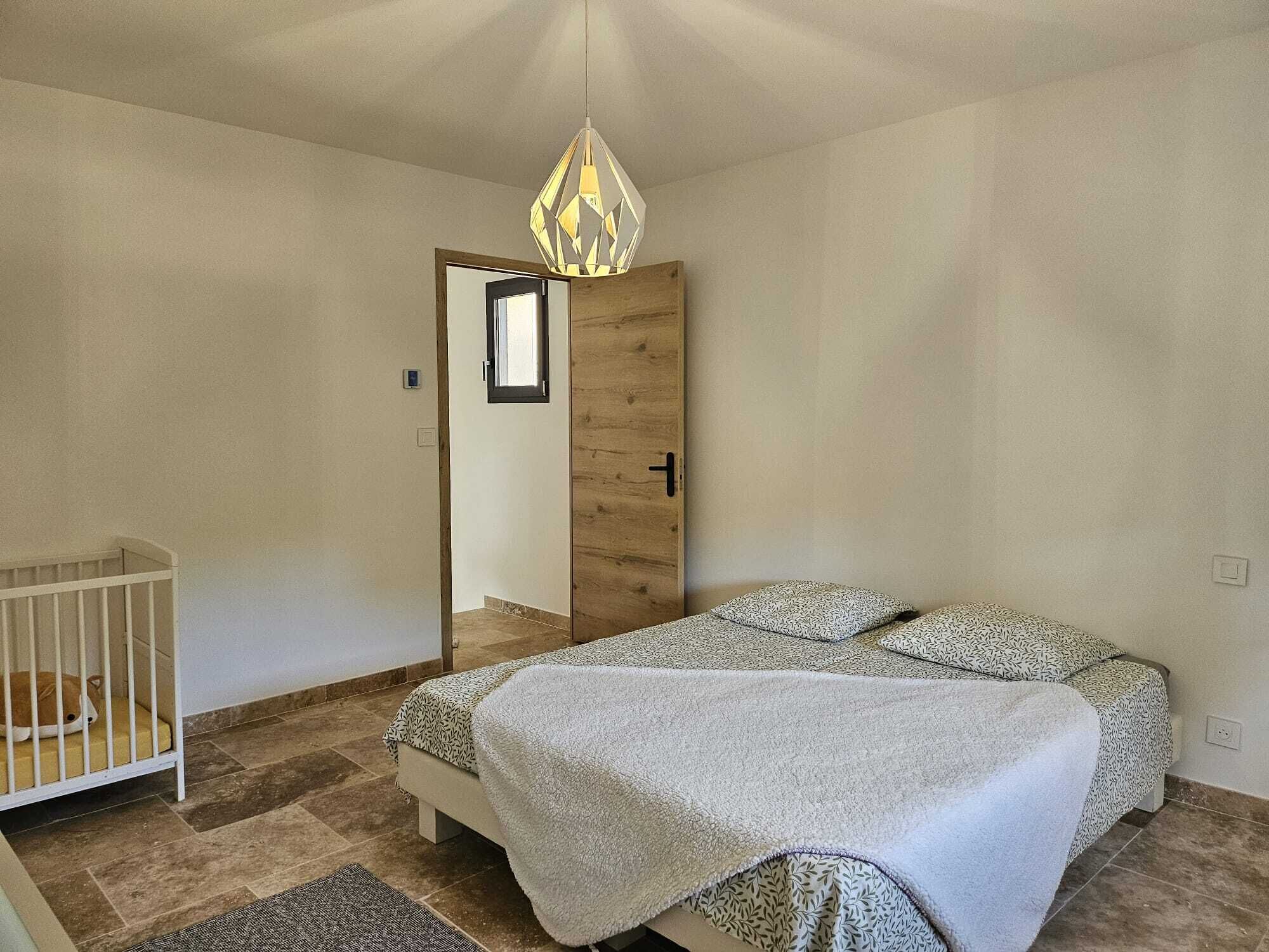 A bedroom with a bed, a crib, a wooden door, and a hanging light fixture.