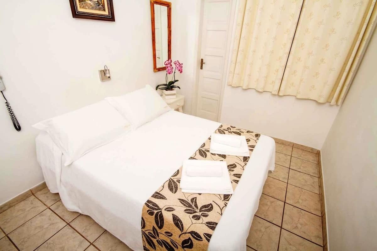 A hotel room with a bed, a bedside table with a phone, a mirror, a door, and a window with curtains.