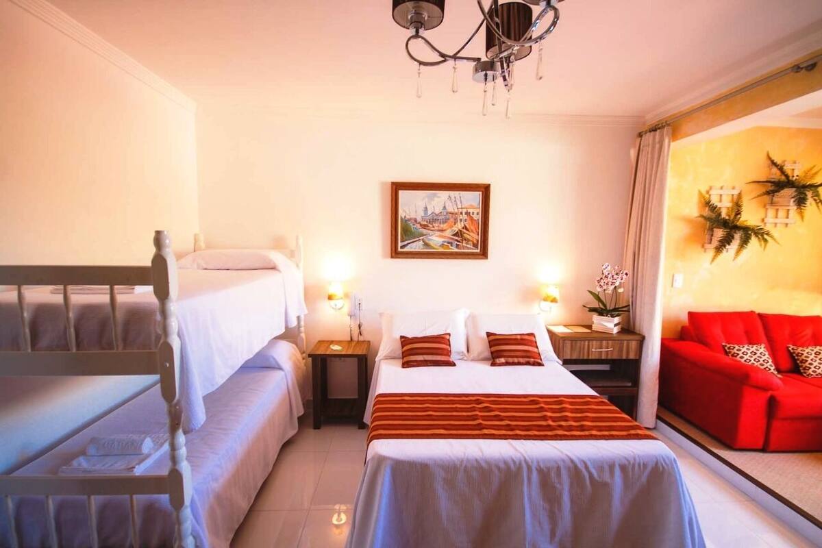 A hotel room with a bunk bed, a single bed, a red sofa, a chandelier, a painting, and a window with curtains.