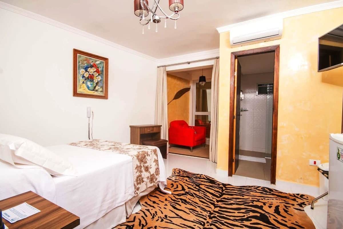 A hotel room with a bed, a red chair, a shower area, and a painting on the wall.