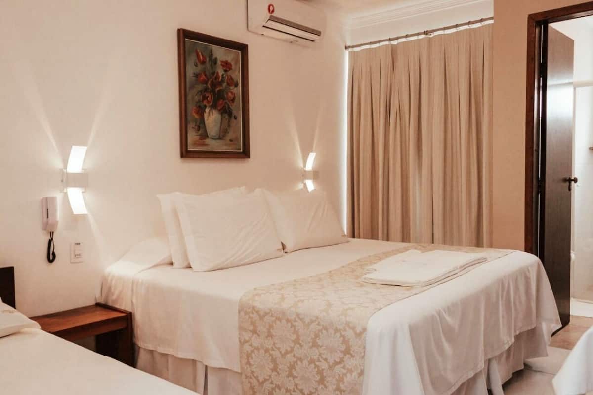 A hotel room with a bed, bedside tables, a painting, an air conditioning unit, and a door.