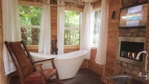 Ein Holzhäuschen im Inneren mit einer Badewanne, einem Esstisch, einem Schaukelstuhl, einem Kamin aus Stein und einem an der Wand montierten Fernseher.