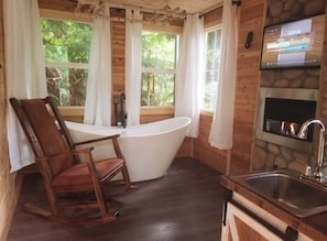 A wooden cabin interior with a bathtub, a dining table, a rocking chair, a stone fireplace, and a TV mounted on the wall.