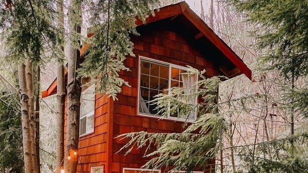 Eine zweistöckige Holzhütte mit einer Holzterrasse und Lichterketten, umgeben von Bäumen.