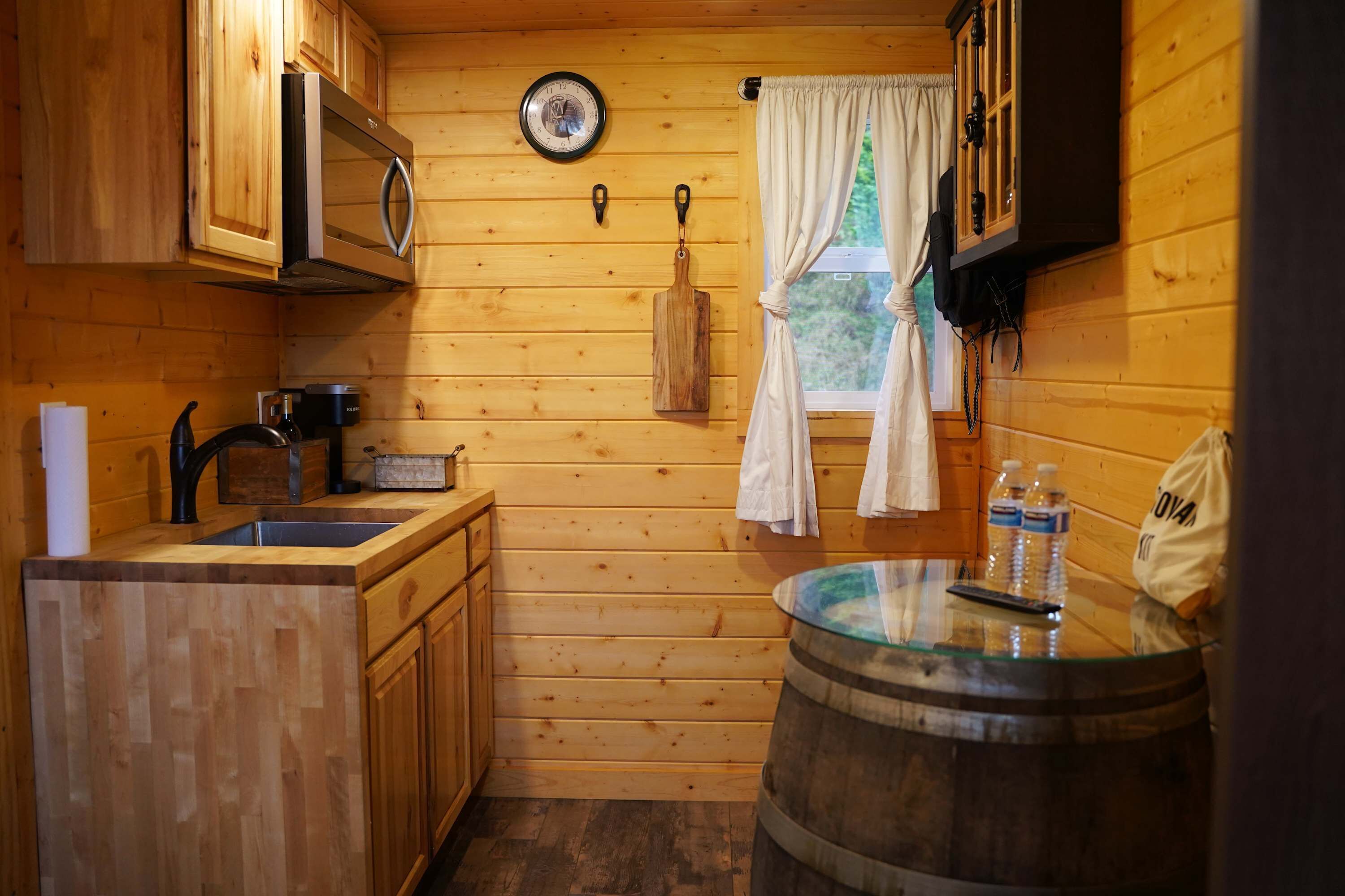 A compact kitchen with wooden cabinetry, a microwave, a sink, and a window with curtains.