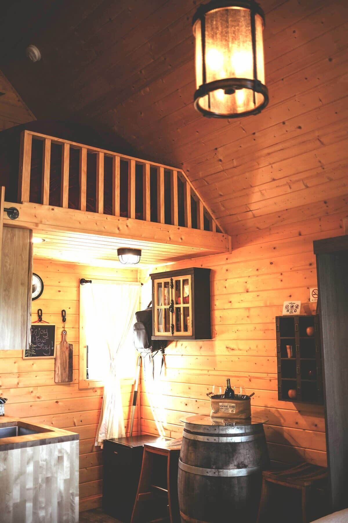 A rustic cabin interior with a wooden staircase, a central stove, and a hanging light fixture.