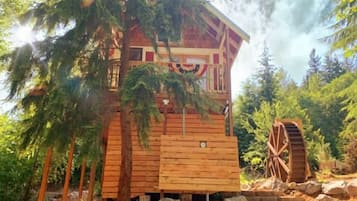A wooden cabin with a balcony, surrounded by trees and a water wheel.