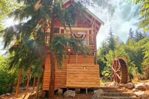 Een houten hut met een balkon, omringd door bomen en een watermolen.